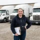 Female small fleet owner standing in front of three white semi-trucks at Columbus Ohio trucking yard holding fleet insurance documents