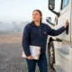 A female owner-operator standing beside her white semi-truck at a Tennessee truck stop in the morning holding commercial truck insurance documents